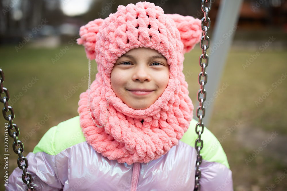 Little funny girl in funny pink hat swinging on swing in park outdoors