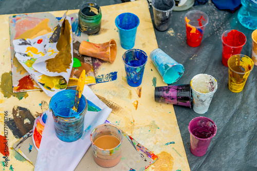Cups filled with colorful paint scattered on a messy table with brushes and paper. Concept of creativity, art, artistic process