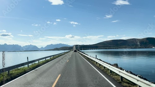 Vehicles are approaching a bridge on a scenic coastal road with mountains in Lofoten islands, Norway