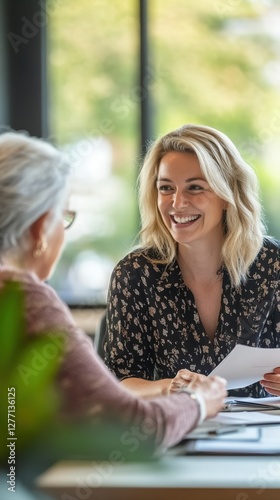 A social worker with light blonde hair smiles warmly while discussing important matters with an elderly client in a bright office. The atmosphere is friendly and supportive