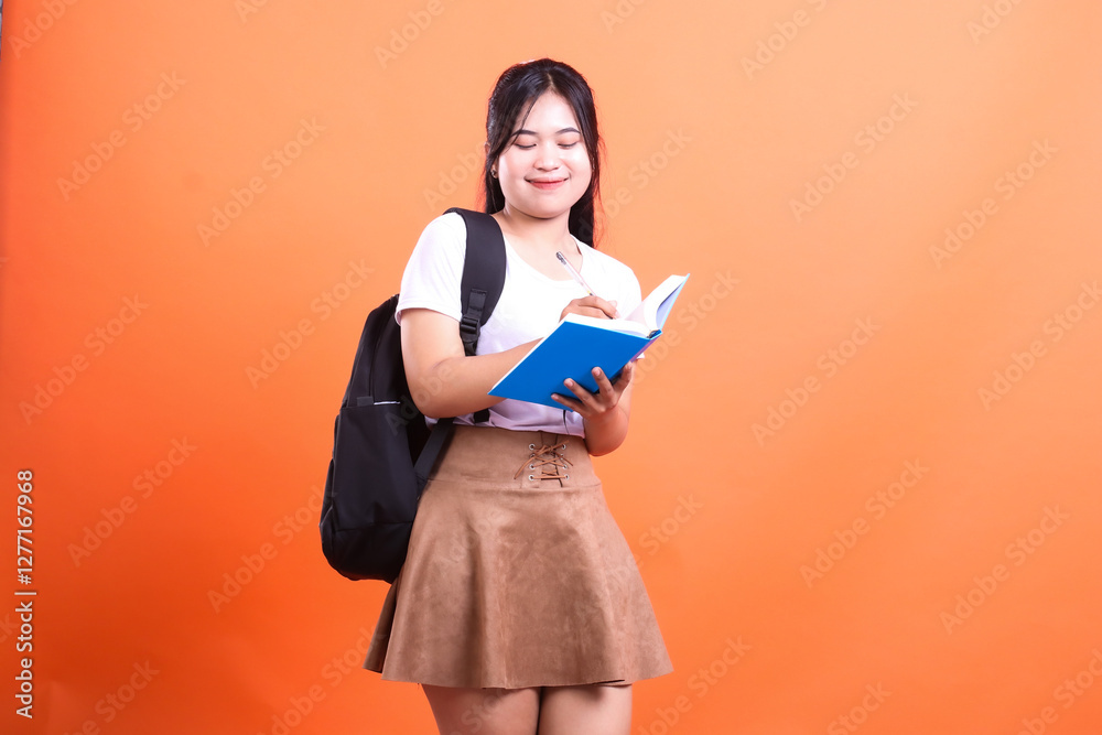 Fototapeta premium A woman with a backpack, wearing a white shirt and a brown skirt, writing in a blue notebook on orange background