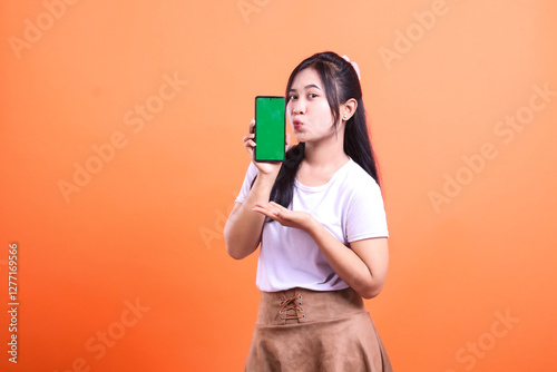 A young woman presents a smart phone cellphone with a green screen, isolated on orange background. She is holding the phone in her hand and her lips are puckered as if kissing