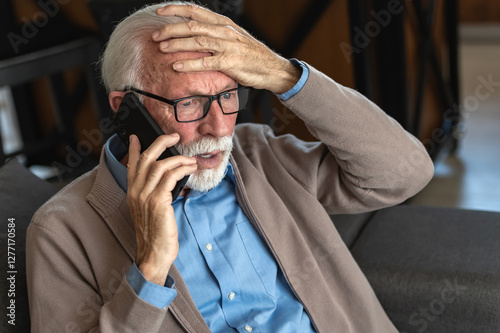 An elderly man with glasses and a beard, wearing a blue shirt and beige cardigan, looks distressed while talking on the phone, holding his head in concern