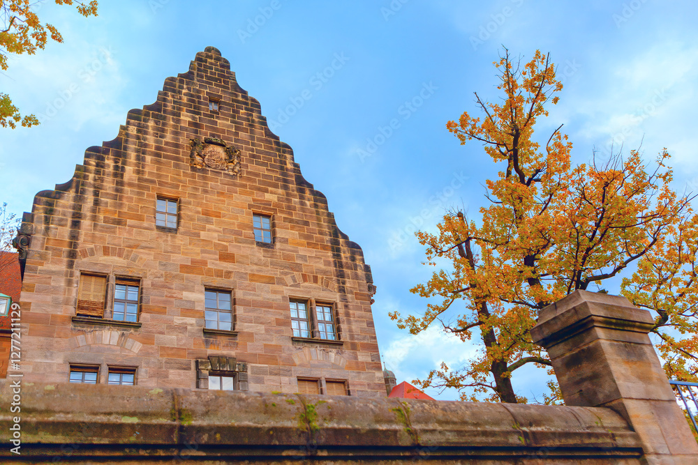 Historic building with stepped gable roof made of stone bricks with windows and a small decorative element near top. Building is framed by autumn trees and a stone wall creating a picturesque scene