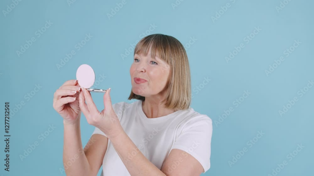 Mature Woman applying makeup in front of mirror against a blue background, showcasing beauty routine and self-care practices in a bright, cheerful setting Mature Woman applying makeup in front of mirror against a blue background, showcasing beauty routine and self-care practices in a bright, cheerful setting