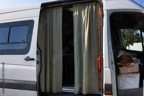 Side view of a camper van with open doors,  beige curtains and a resting traveler, capturing a cozy van life moment, freedom, and a nomadic lifestyle on the road.

