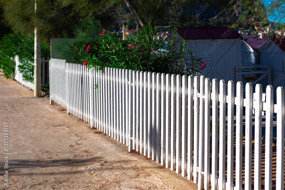 White picket fence running alongside a dirt path, with green bushes and pink flowers growing behind fence. In background, there are white buildings with red roofs partially visible through the foliage