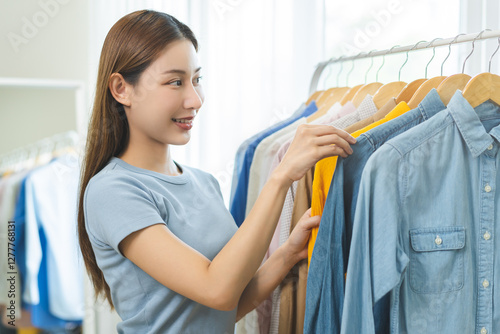 Choose clothes to wear, attractive asian young woman, girl try on appare, happy choice of shirt, female outfit on hanger in wardrobe in room closet at home. Deciding blouse what to put on which one photo