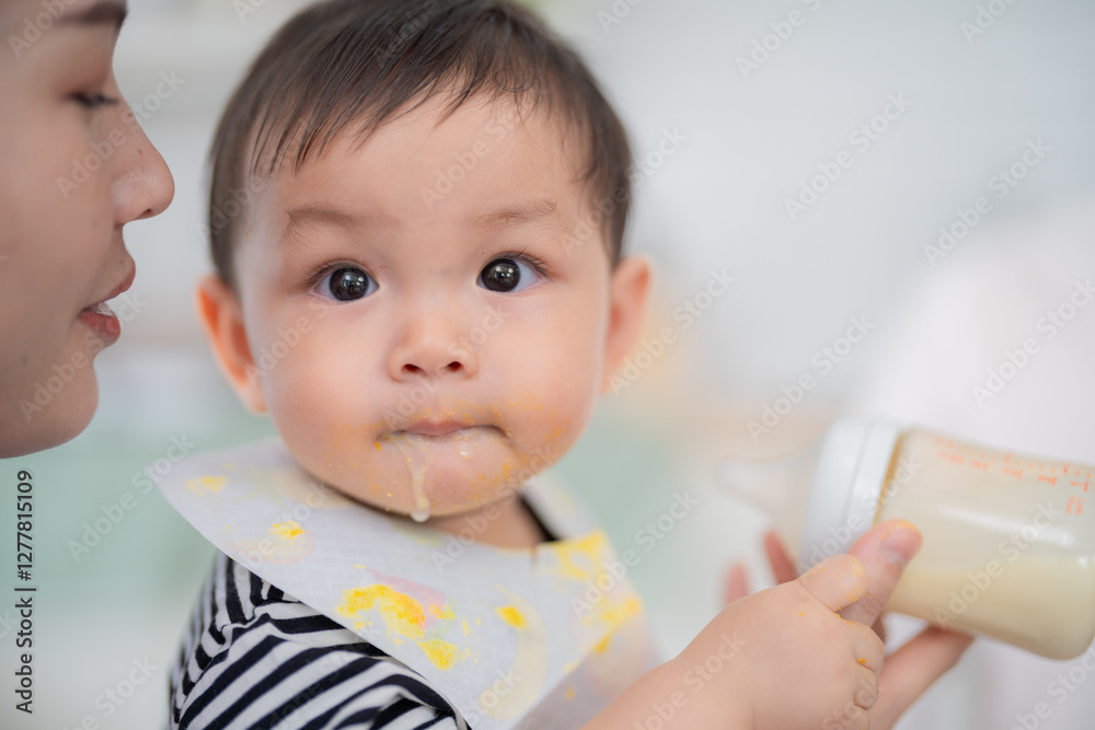 Child enjoying meal while holding bottle in kitchen during daytime with caregiver close by Child enjoying meal while holding bottle in kitchen during daytime with caregiver close by
