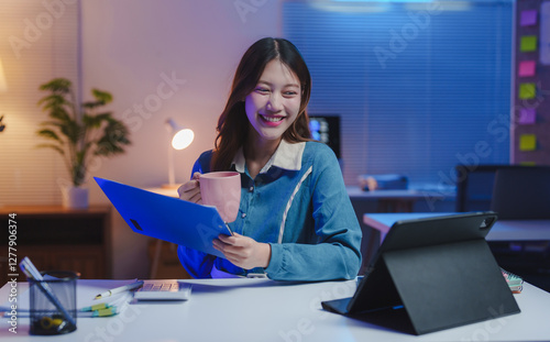 Young, happy asian businesswoman enjoying coffee while reviewing documents and working late at night in the office, utilizing a tablet computer for her tasks and projects