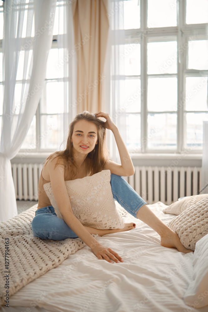 Young woman sitting on bed and enjoying natural light in cozy bedroom during morning Young woman sitting on bed and enjoying natural light in cozy bedroom during morning