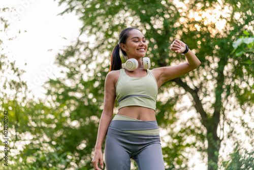 Young Asian woman enjoying music on headphones while exercising in green park. Sporty woman using headphone in outdoor nature setting.
