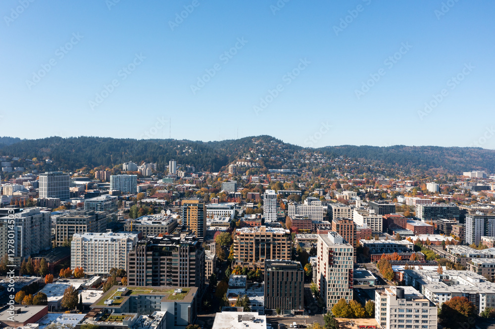 Fototapeta premium Aerial drone view of downtown Portland, Oregon, featuring modern high-rises, urban architecture, and tree-lined streets with Forest Park and the West Hills in the background on a clear autumn day.