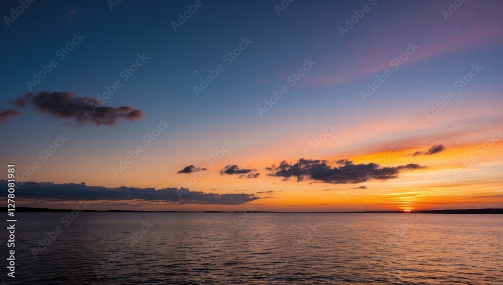Obraz premium A serene sunset over a calm sea, vibrant orange and yellow hues blending into a soft blue sky, distant dark coastline, gentle waves reflecting the colourful light