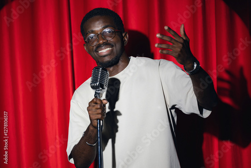 Concept comedian show open mike. Happy young african american man comic performing standup monologue on stage with red curtains. photo