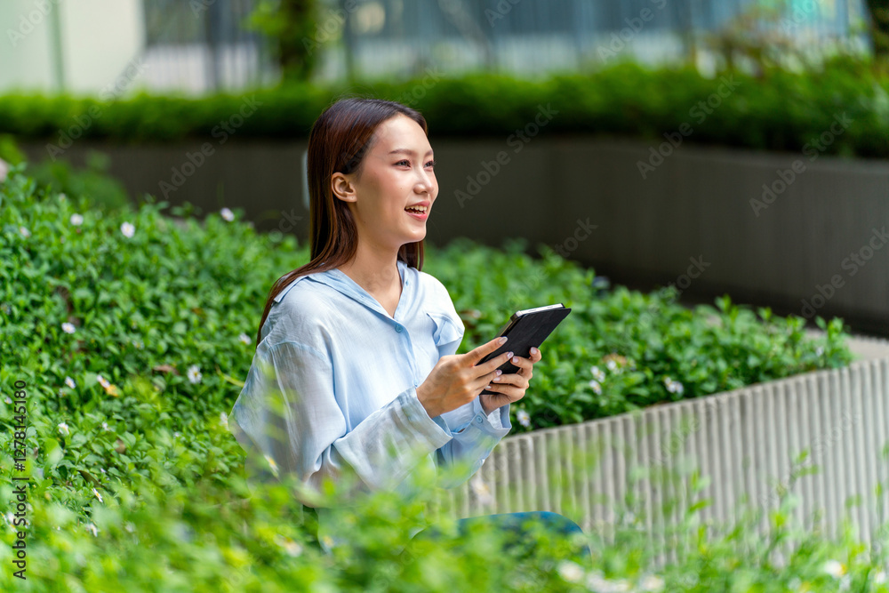 Fototapeta premium Young Asian Businesswoman In Casual Business Outfit Using Tablet Outdoors Sitting In Front a Modern Office Building