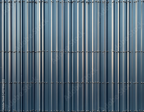 Exterior view of building with corrugated metal panels. Cladding dark gray color, scalloped design. Horizontal stripes, shadows present on surface. Metal sheets rivets. Structure looks modern,