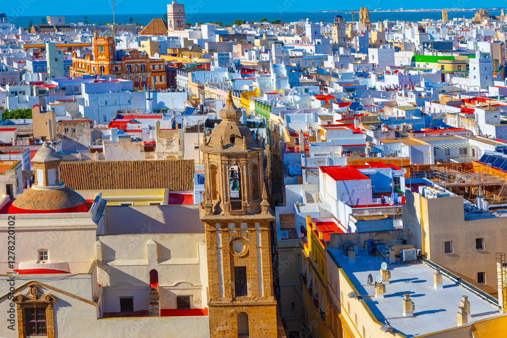 Aerial view of city of Cadiz, Spain, dense array of white buildings with flat roofs and prominent church tower in foreground. Vibrant colors of the rooftops and the visible coastline highlight Cadiz