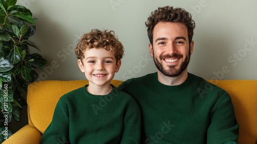 A smiling father and son sit together on a cozy couch, both wearing matching green sweaters, surrounded by indoor plants.