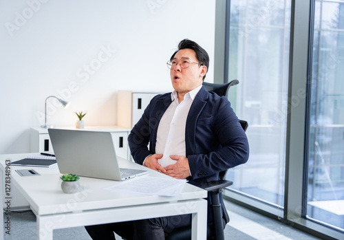 Ill Asian businessman seated at a modern office desk, showing signs of abdominal discomfort, wearing a business suit with glasses, surrounded by minimalistic workspace characterized by natural light.