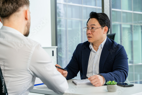 Asian businessman in a stylish office meeting with individual, offering insights, wearing business attire communicative interaction highlighting confidence and expertise in a corporate environment.