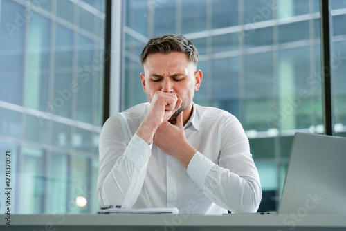 Professional businessman sitting in a bright office, wearing a white shirt, feeling stressed and deep in thought, surrounded by modern glass architecture conveying workplace tension.