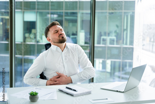 Professional office worker in a bright modern office, seemingly struggling with stomach discomfort, seated at a desk with laptop and documents, conveying an emotion of unease or stress.