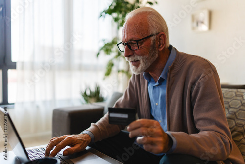 A cheerful elderly man with a beard sits comfortably on a sofa in a bright, naturally lit room, uses a laptop while holding a credit card, smiling as he shops online or makes a secure digital paymet photo