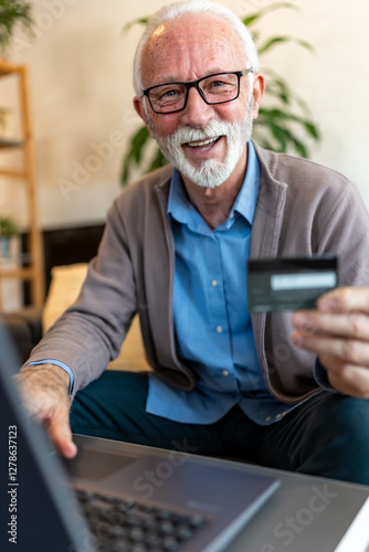 A cheerful elderly man with a beard sits comfortably on a sofa in a bright, naturally lit room, uses a laptop while holding a credit card, smiling as he shops online or makes a secure digital paymet photo