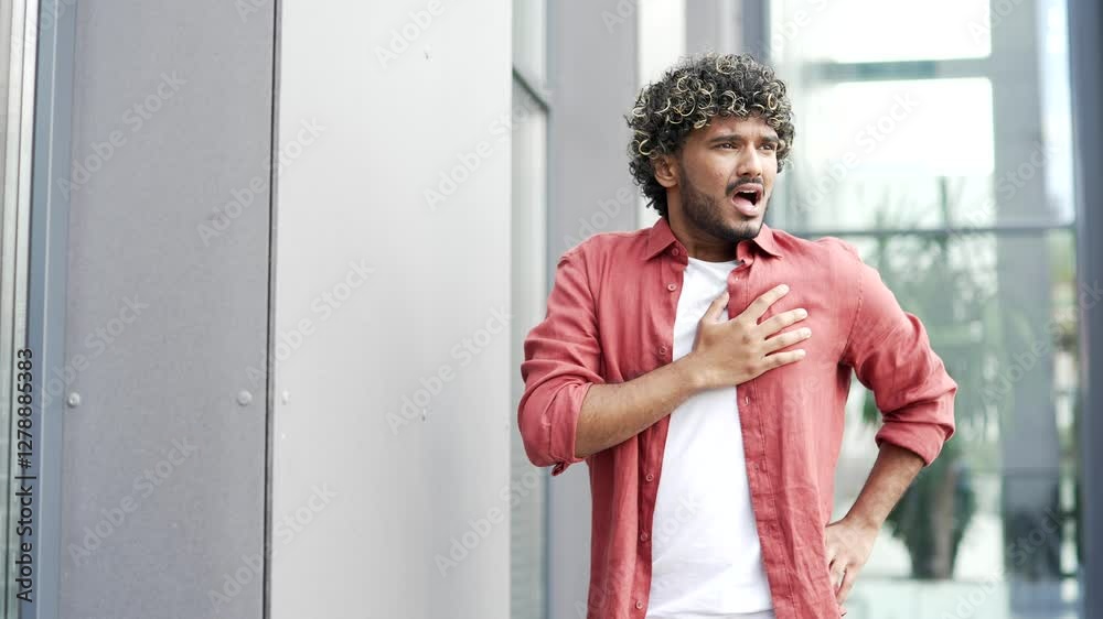 Sick young businessman has a heart attack while standing on the street near modern office building. Upset handsome male suffers from tension in the chest and holds a sore spot with his hands. Close up