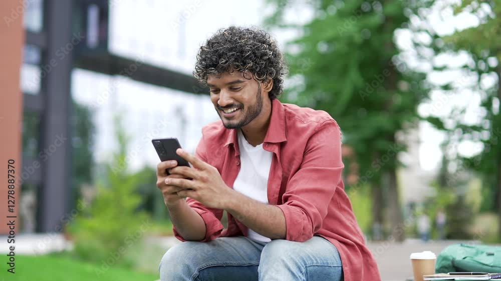 Young smiling man uses a mobile phone while sitting on a bench on street near a modern office building. Cheerful handsome male in a red shirt chats online with friend or browses social media. Close up Young smiling man uses a mobile phone while sitting on a bench on street near a modern office building. Cheerful handsome male in a red shirt chats online with friend or browses social media. Close up