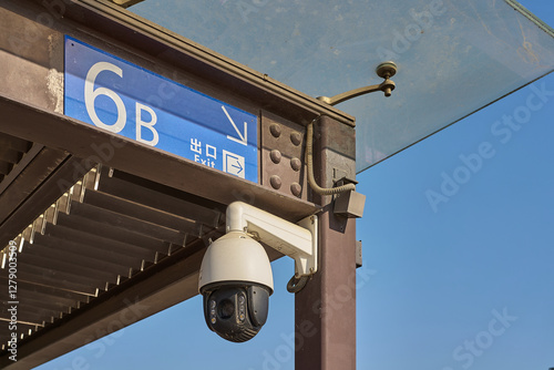 Security camera monitoring area near exit sign on a sunny day, with a blue sky backdrop and metal frame.