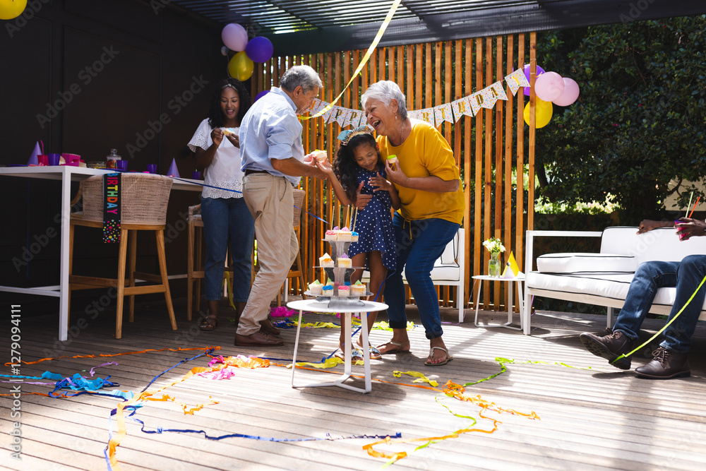 Fototapeta premium Celebrating birthday in garden, diverse family dancing with child, grandparents smiling joyfully