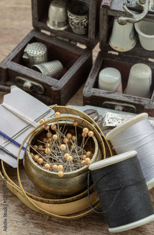 Sewing needles stuck in a white paper, Thimbles in small wooden treasure chest, Spools of black and white thread, Measuring tape and Steel scissors including pins in brass cup on wooden background.