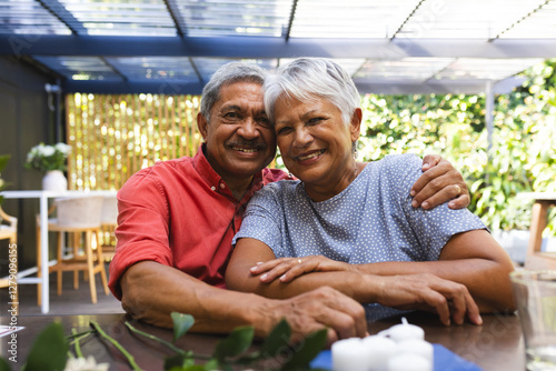 Elderly diverse couple sitting together in garden, smiling and embracing at diverse family gathering