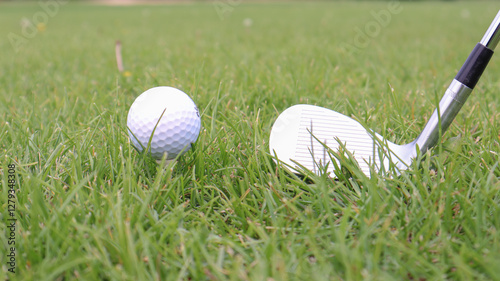 Close-up on a golf ball set on a tee beside a golf club in lush green grass, conveying preparation to start the game. Suitable for themes related to sports, recreation, golf, or outdoor activities.