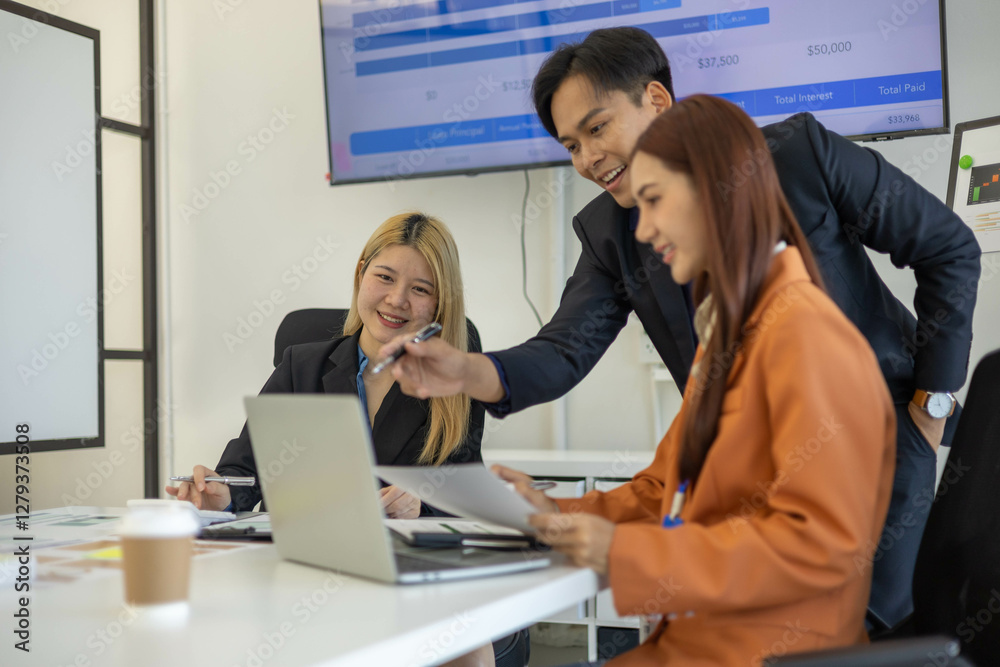 Three people are sitting at a table with a laptop open. One of them is laughing while the other two look on. The man in the middle is giving a presentation to the other two

