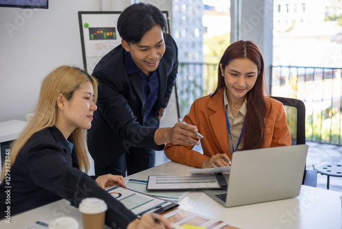 Three people are sitting at a table with a laptop open. One of them is laughing while the other two look on. The man in the middle is giving a presentation to the other two


