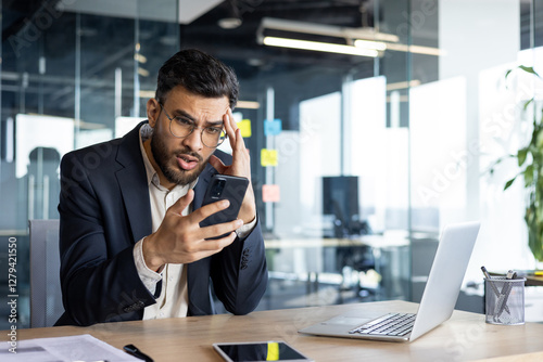 Upset and disappointed man with phone in hands inside office at workplace, businessman reading bad news