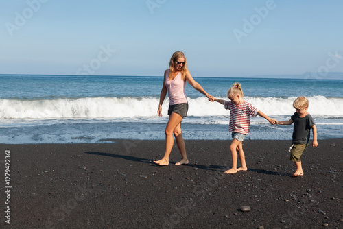 Happy family - young mother, daughter, baby son hold hands, walk together by water pool along sea surf on black sand beach. Travel, active lifestyle, parents with child on summer vacation