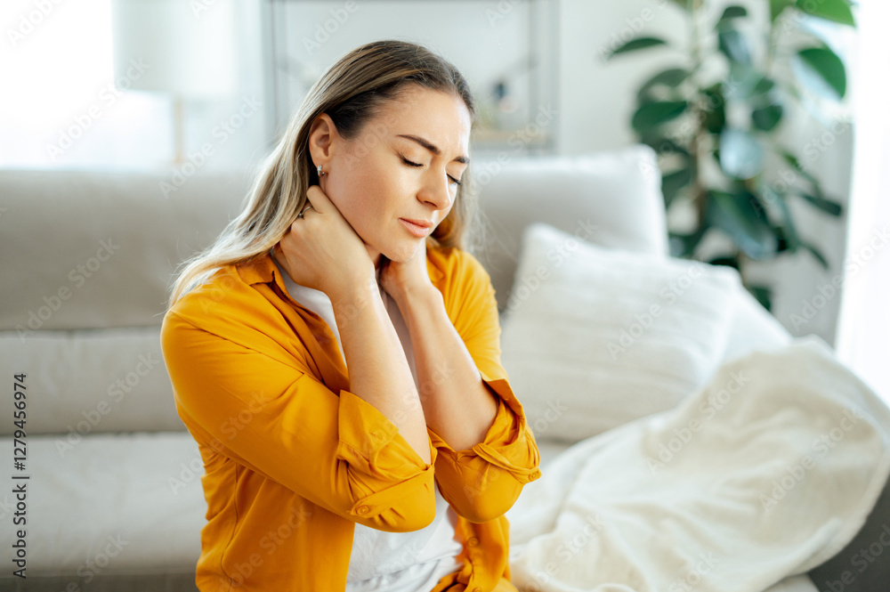 Concept of neck pain, stiffness, or muscle tension. Caucasian exhausted woman in a yellow shirt sitting on a cozy couch at home, holding her neck with a pained facial expression Concept of neck pain, stiffness, or muscle tension. Caucasian exhausted woman in a yellow shirt sitting on a cozy couch at home, holding her neck with a pained facial expression