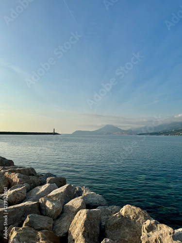 Vertical view of Bar, Montenegro, in warm sunset light with clear blue sky snd seascape, a distant horizon line, a lighthouse, and mountains in the background