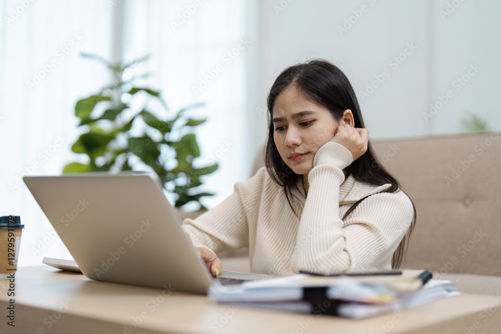Young Asian woman looking concerned while working on a laptop at home, reflecting on challenges. Young Asian woman looking concerned while working on a laptop at home, reflecting on challenges.