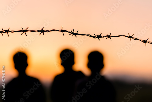 Silhouetted figures stand behind barbed wire against a soft sunset sky. The scene evokes themes of restriction, separation, and human struggles.