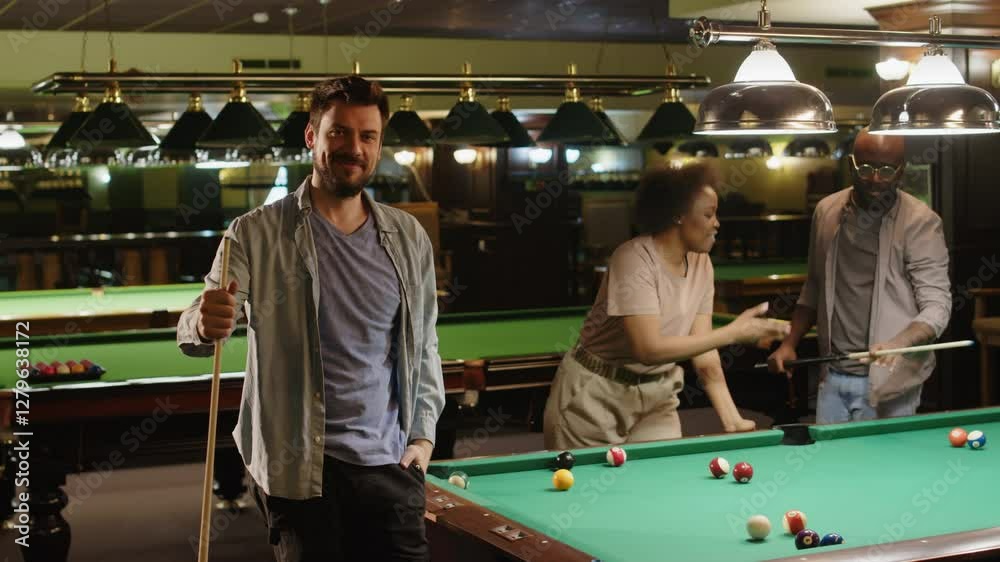 Full medium portrait of young bearded Caucasian man posing by baize table in billiard club with cue stick in hand while playing with diverse friends in evening, look at camera