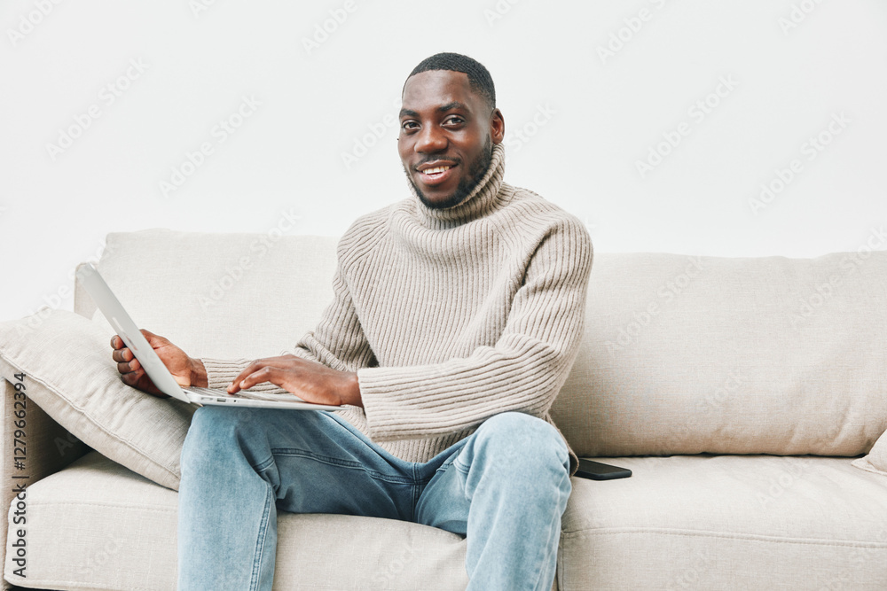 Smiling young Black man in a cozy beige turtleneck sweater working on a laptop at home, creating a relaxed and productive atmosphere Smiling young Black man in a cozy beige turtleneck sweater working on a laptop at home, creating a relaxed and productive atmosphere