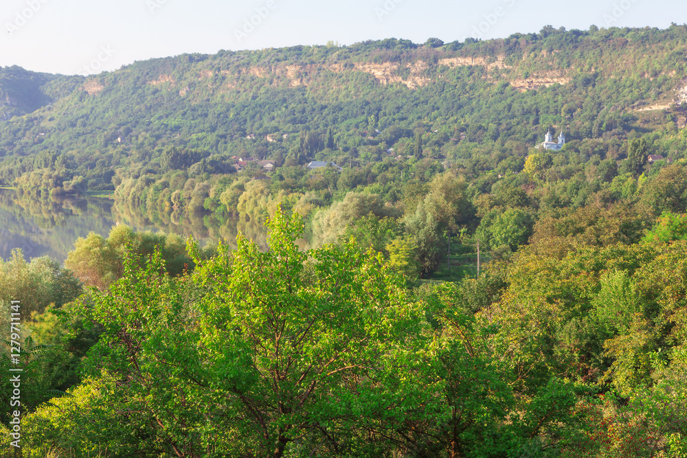 Scenic landscape in Moldova with lush green forests, a calm river, and rolling hills. Distant church adds charm to serene countryside, bathed in warm sunlight, creating peaceful and natural atmosphere