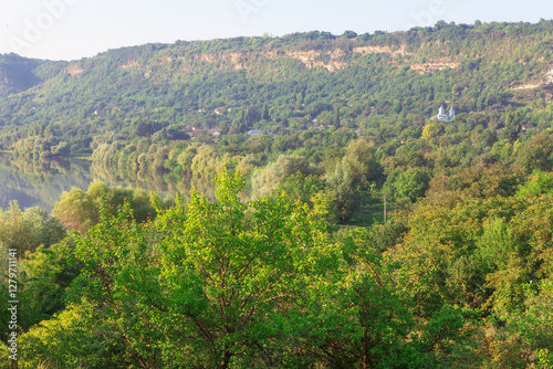 Obraz na płótnie Scenic landscape in Moldova with lush green forests, a calm river, and rolling hills. Distant church adds charm to serene countryside, bathed in warm sunlight, creating peaceful and natural atmosphere w salonie