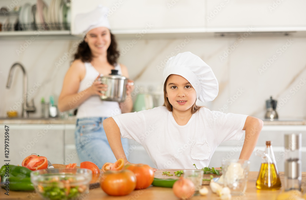 Happy little girl posing by kitchen-table while her mother standing behind in the kitchen Happy little girl posing by kitchen-table while her mother standing behind in the kitchen