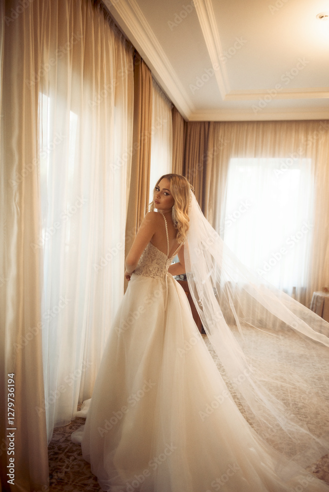 Blonde bride with veil standing near window in white dress with train. Blonde bride with veil standing near window in white dress with train.